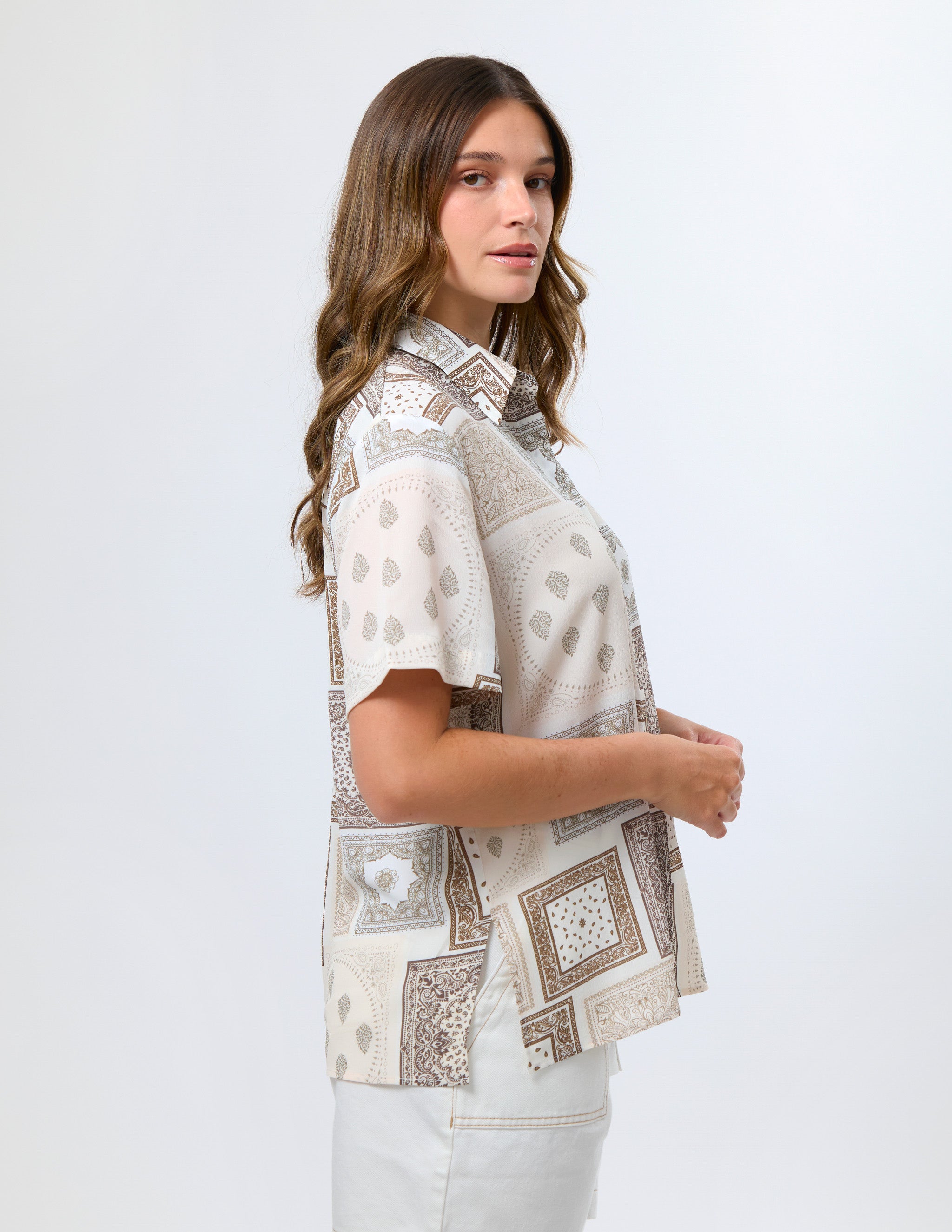A woman wearing a short-sleeve button-up shirt with a beige and brown geometric patchwork print, paired with white pants, photographed against a light gray studio background.