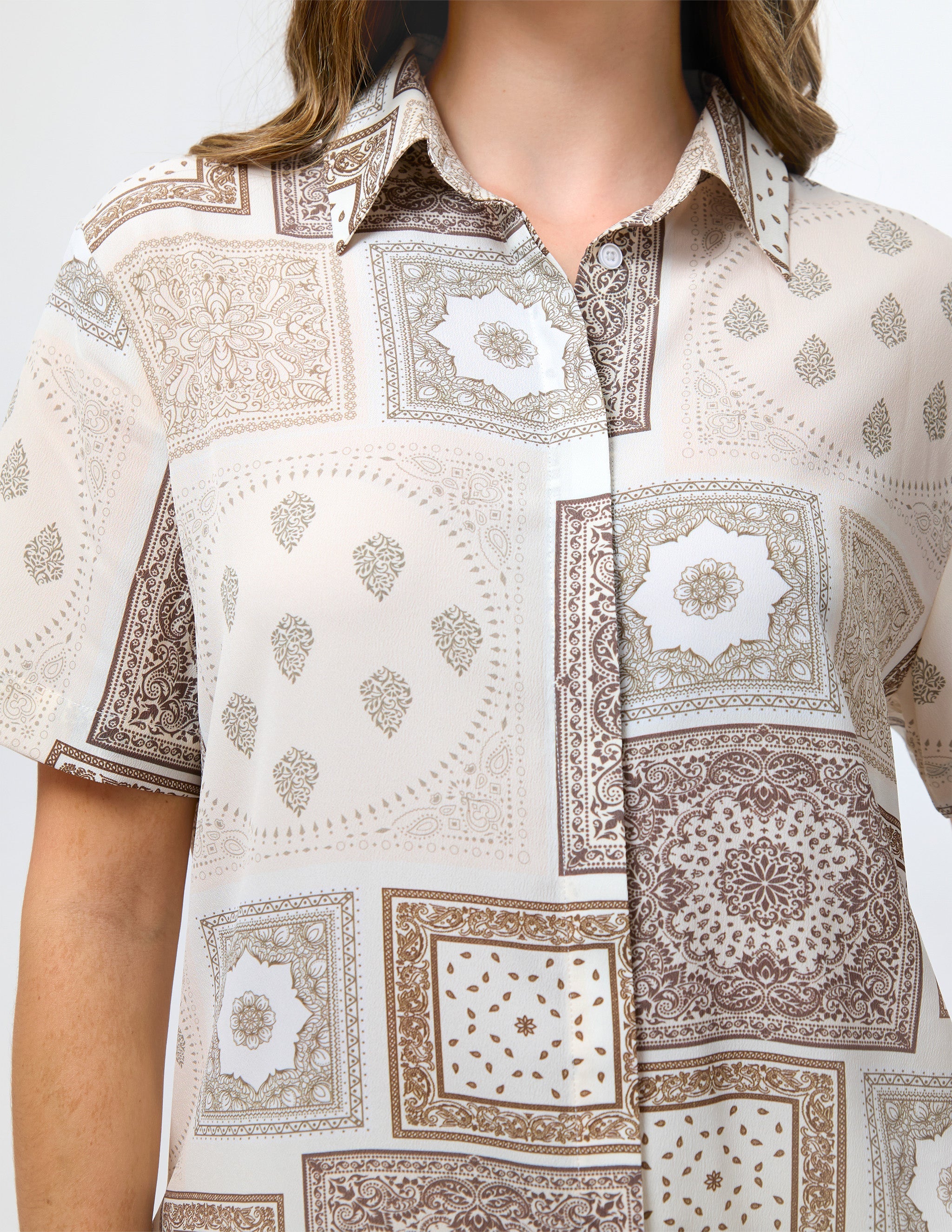 A close-up of a model wearing a short-sleeve button-up shirt with a beige, brown, and white patchwork print featuring geometric and floral motifs.