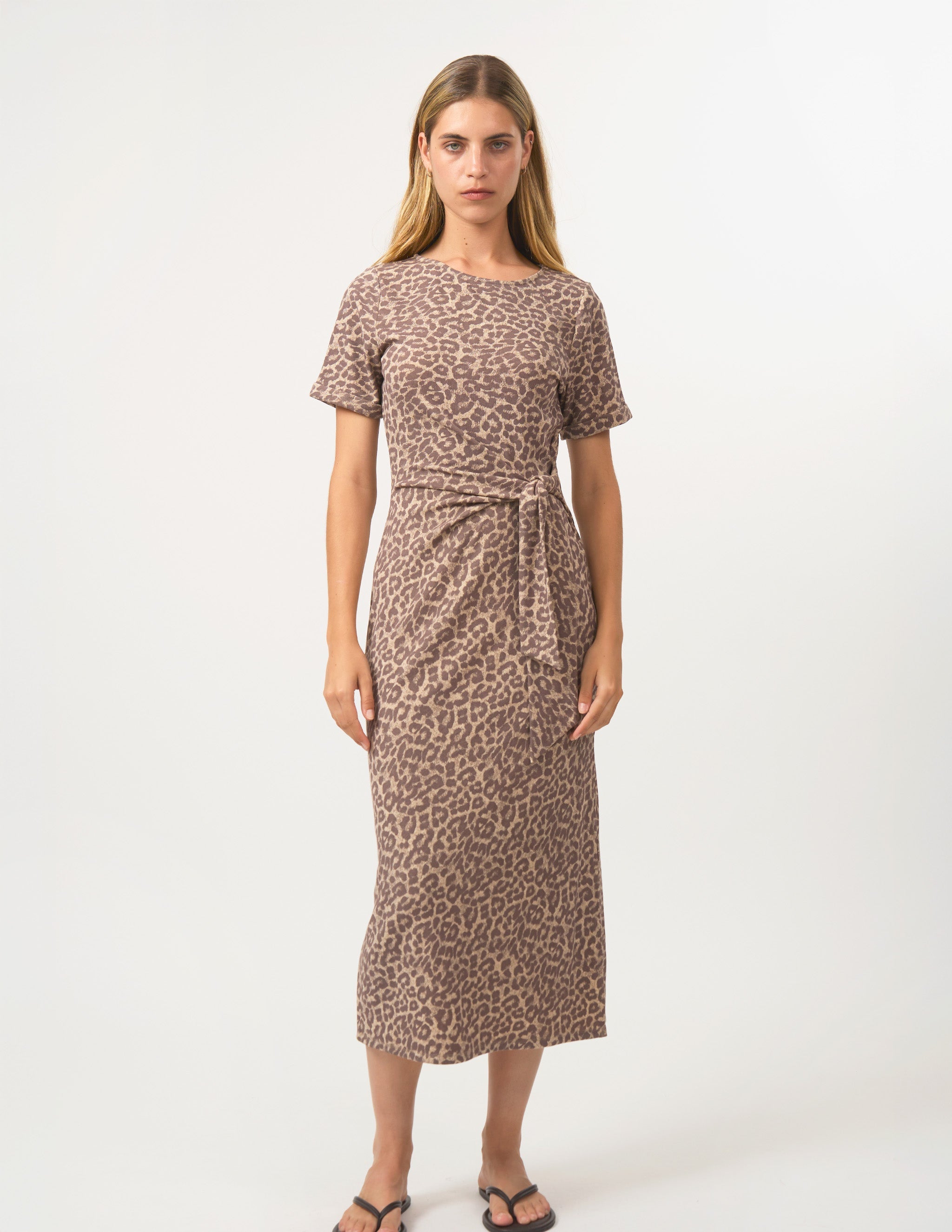 A woman modeling a short-sleeve midi dress with a brown and beige leopard print pattern, featuring a tie waist and side pockets, set against a plain white studio background.
