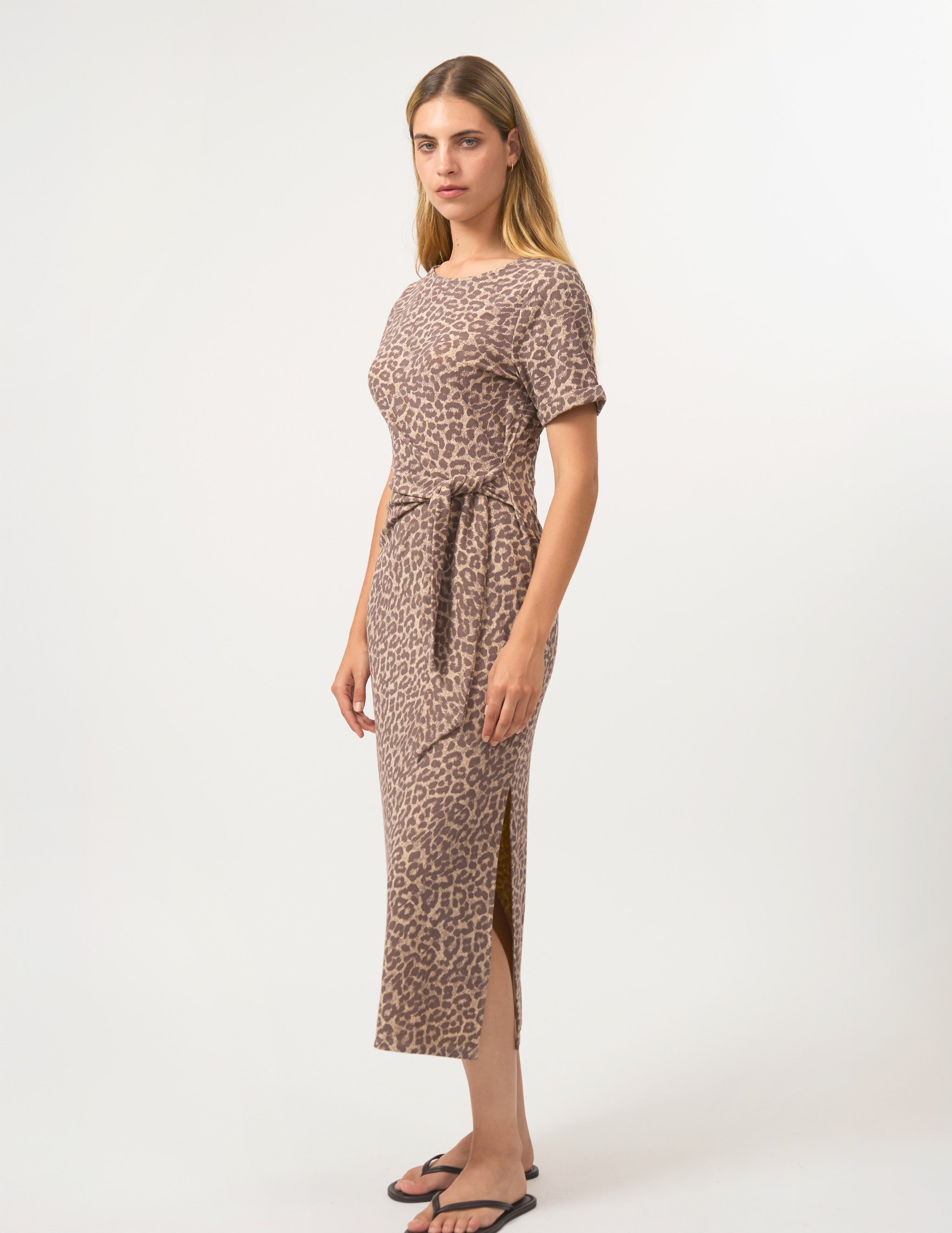 A woman modeling a short-sleeve midi dress with a brown and beige leopard print pattern, featuring a tie-waist belt and side slits, set against a plain white studio background.