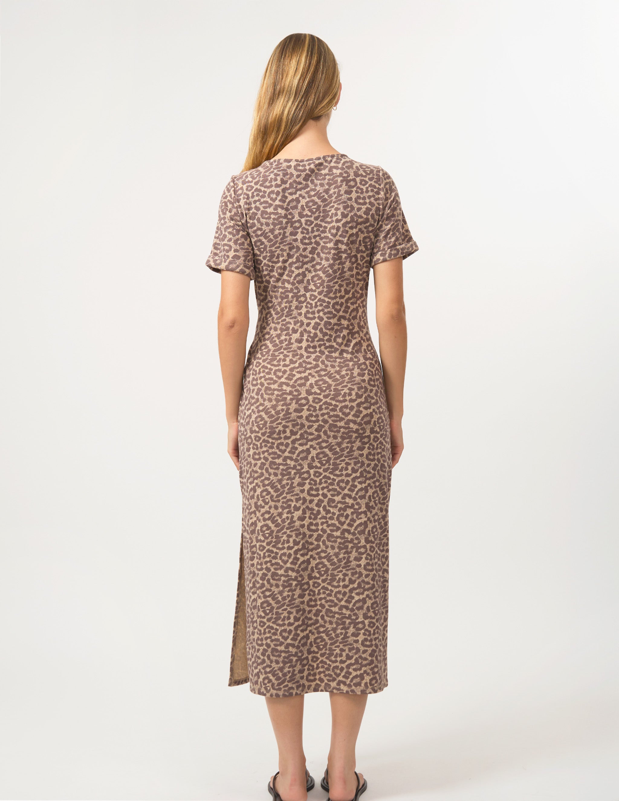 A woman viewed from the back wearing a short-sleeve leopard-print midi dress with a side slit, standing against a plain light gray studio background.