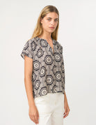 A woman modeling a short-sleeve blouse with a black and off-white circular mandala pattern, paired with white pants, against a plain white studio background.