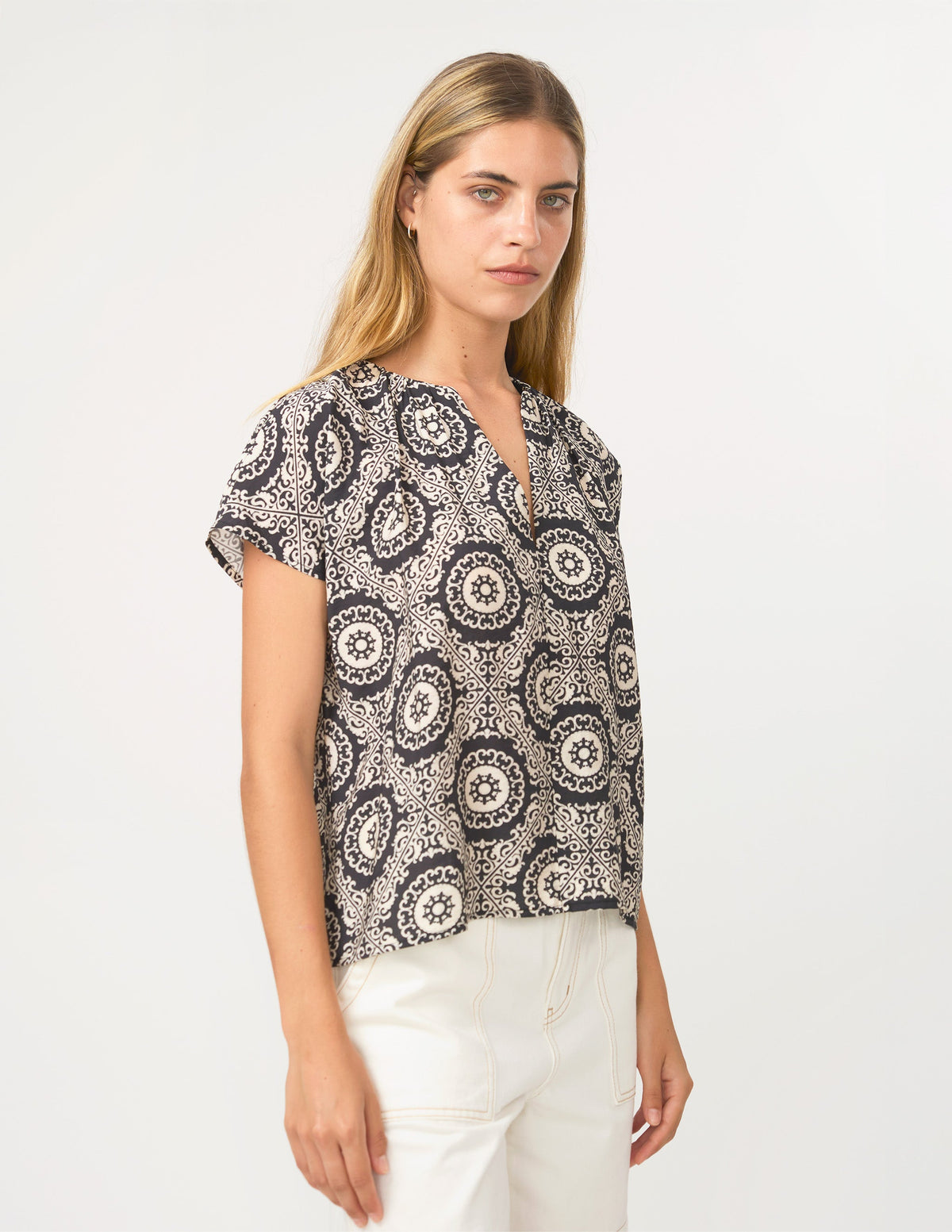 A woman modeling a short-sleeve blouse with a black and off-white circular mandala pattern, paired with white pants, against a plain white studio background.