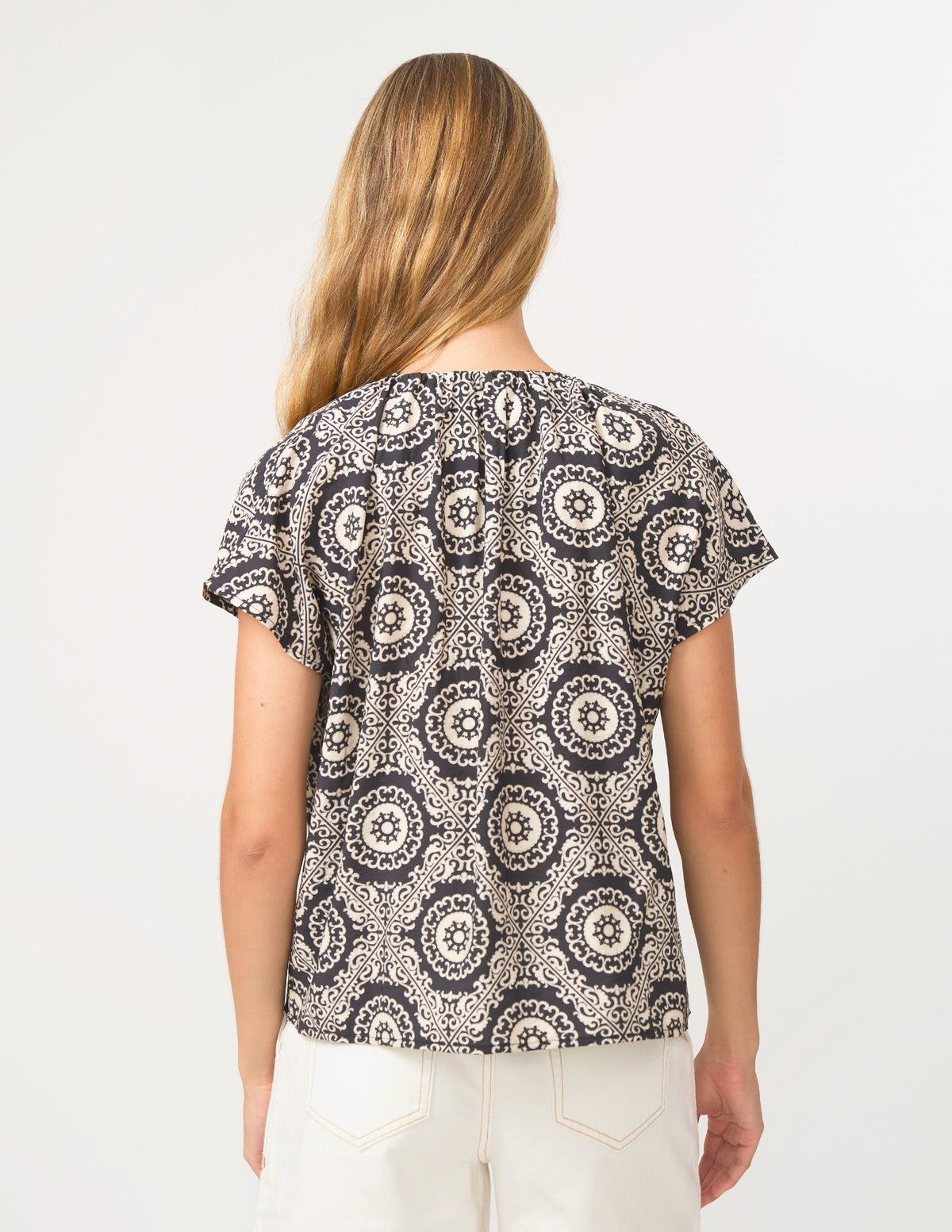 A woman viewed from the back wearing a short-sleeve top with an intricate black, white, and gray circular mandala pattern, paired with off-white pants, against a plain white background.