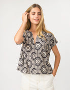 A woman modeling a short-sleeve V-neck blouse with a black and beige circular mandala pattern, paired with white pants, against a plain light gray background.