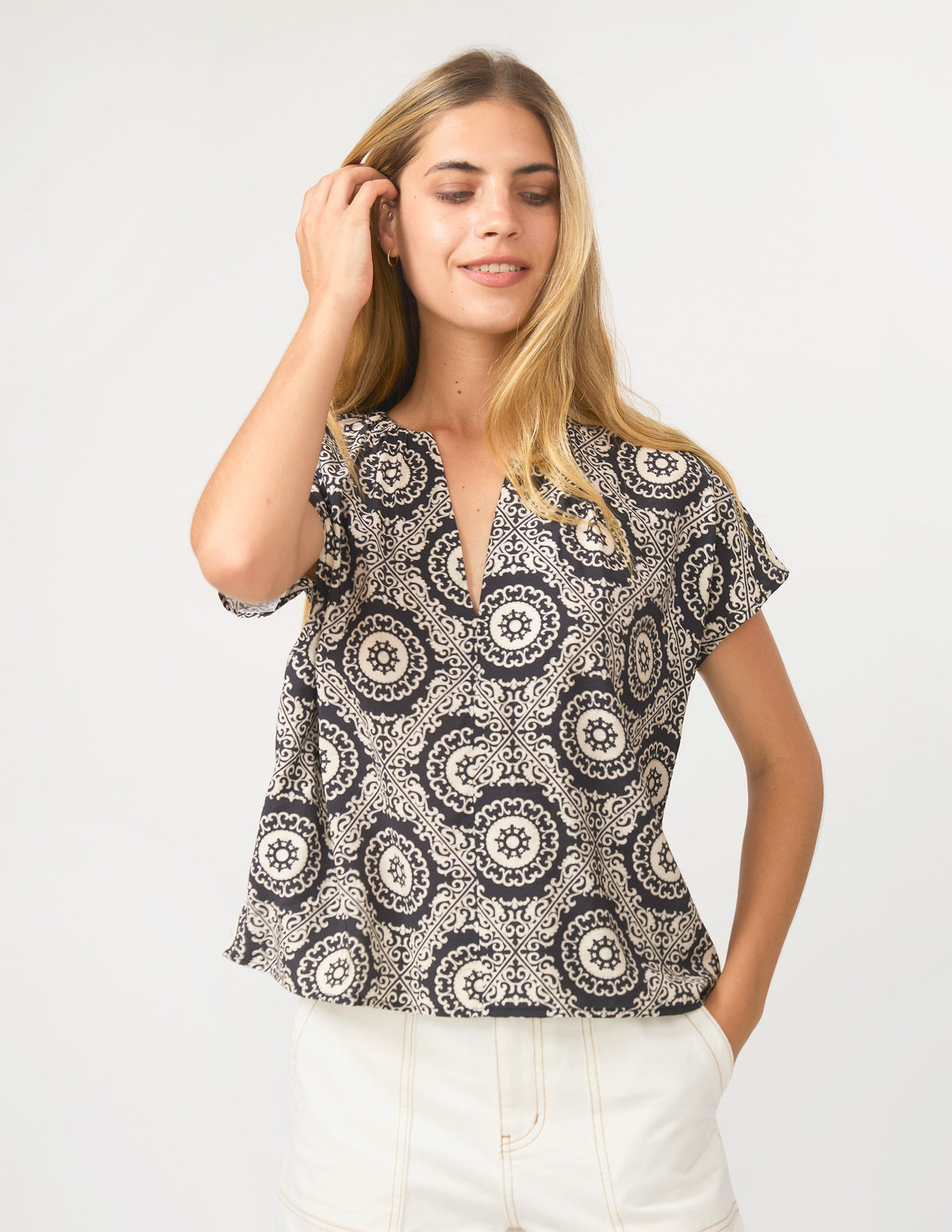 A woman modeling a short-sleeve V-neck blouse with a black and beige circular mandala pattern, paired with white pants, against a plain light gray background.