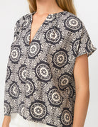 A woman wearing a short-sleeve blouse with a black and off-white bohemian mandala pattern, shown from the chest down against a plain white background.