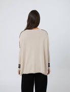 A woman viewed from behind wearing a light beige knit sweater with black stripe details on the shoulders and cuffs, paired with black pants, against a plain light gray background.