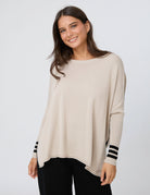 A woman wearing a light beige knit poncho sweater with black and white striped cuffs, paired with black pants, against a light gray studio background.