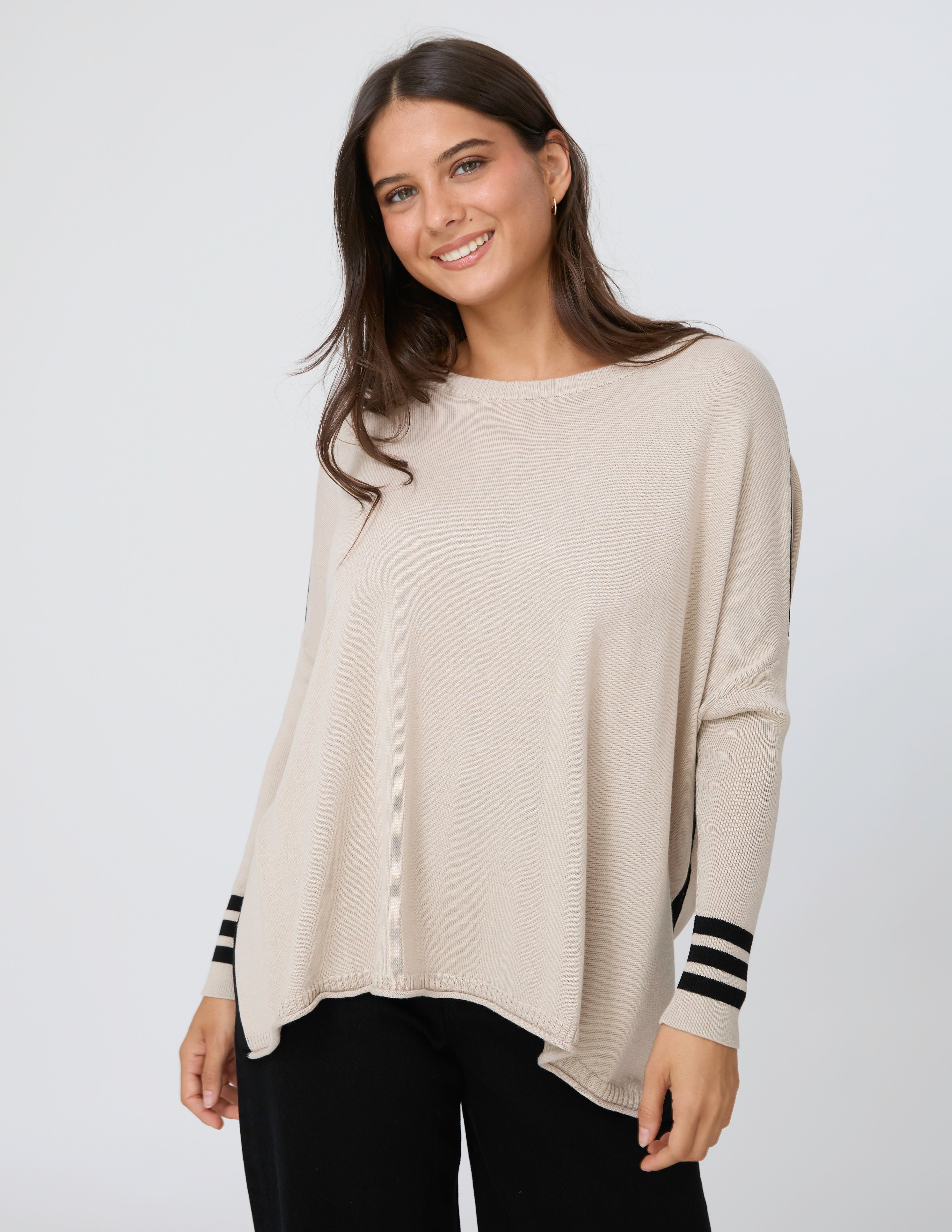 A woman wearing a light beige knit poncho sweater with black and white striped cuffs, paired with black pants, against a light gray studio background.