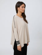 A woman modeling a light beige knit sweater with black stripe details on the sleeve cuffs and a black accent line on the shoulder, paired with black pants, against a plain light gray background.