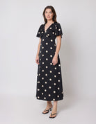 A woman modeling a black midi dress with large beige polka dots, short flutter sleeves, and a V-neckline, photographed against a light gray studio background.
