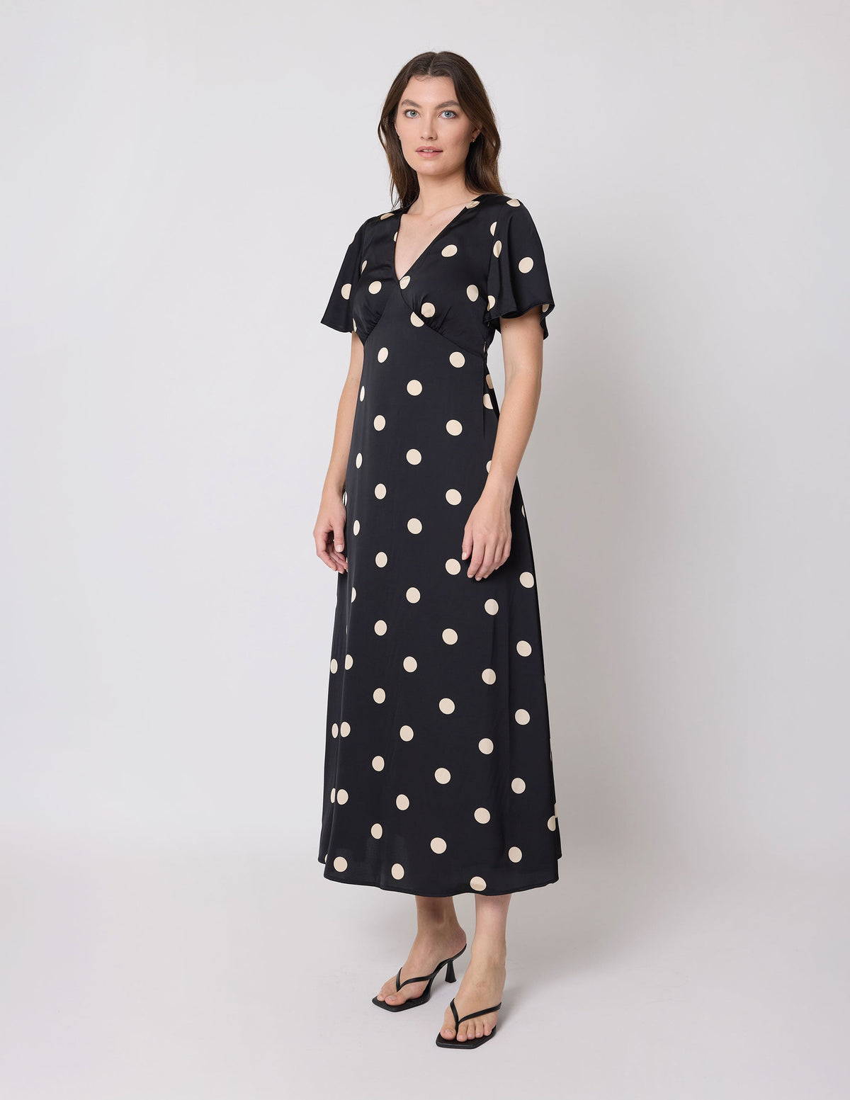 A woman modeling a black midi dress with large beige polka dots, short flutter sleeves, and a V-neckline, photographed against a light gray studio background.