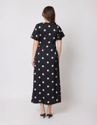 A woman viewed from the back wearing a black midi dress with large beige polka dots, short sleeves, and a round neckline, paired with black pointed-toe heels against a light gray studio background.