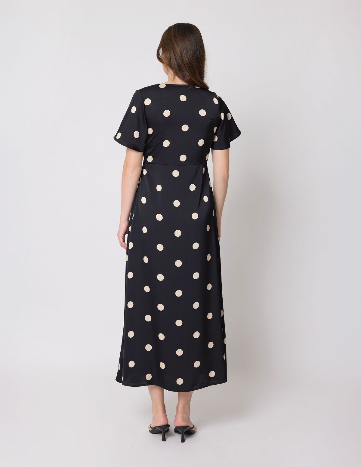 A woman viewed from the back wearing a black midi dress with large beige polka dots, short sleeves, and a round neckline, paired with black pointed-toe heels against a light gray studio background.