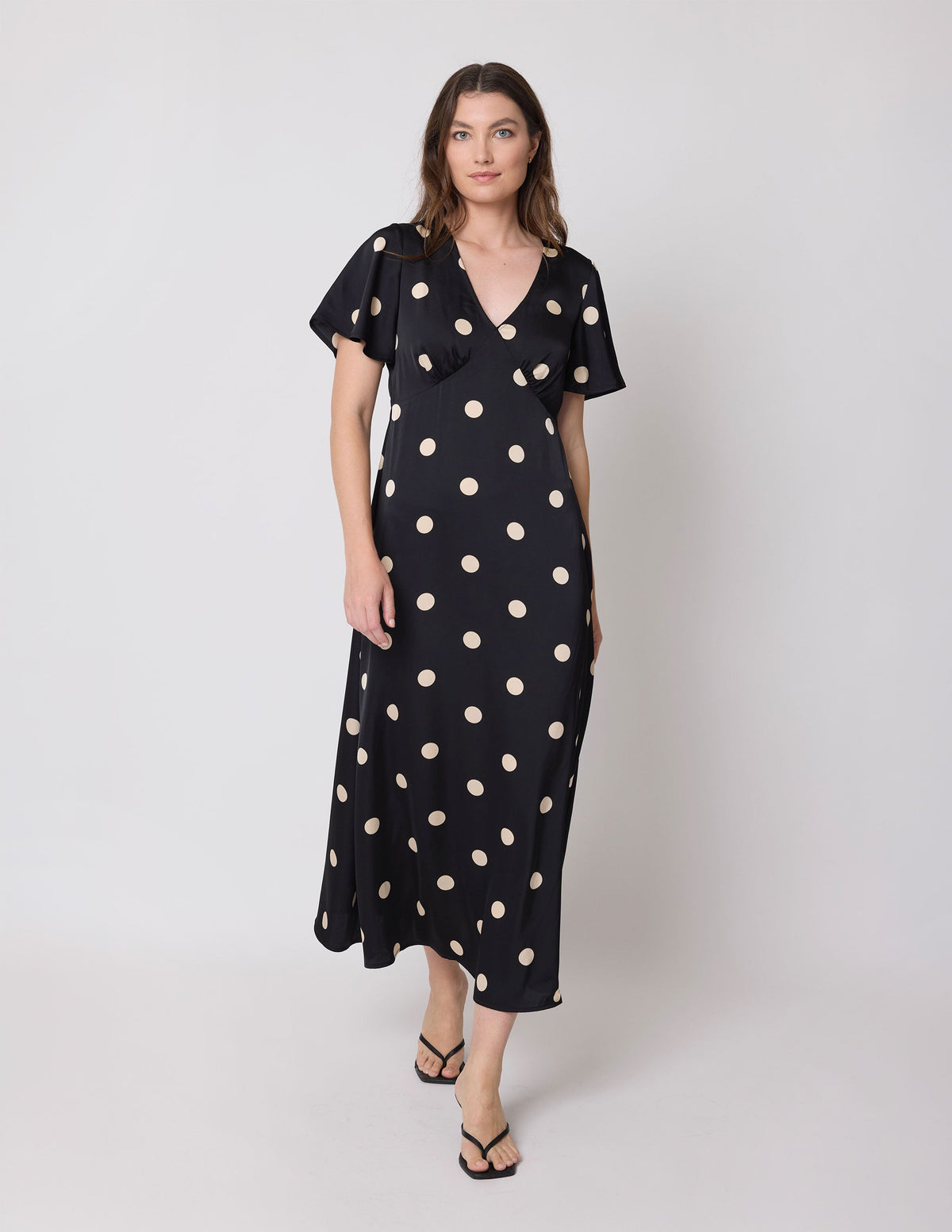 A woman modeling a black midi wrap dress with beige polka dots, flutter sleeves, and a ruffled V-neckline, set against a plain light gray studio background.
