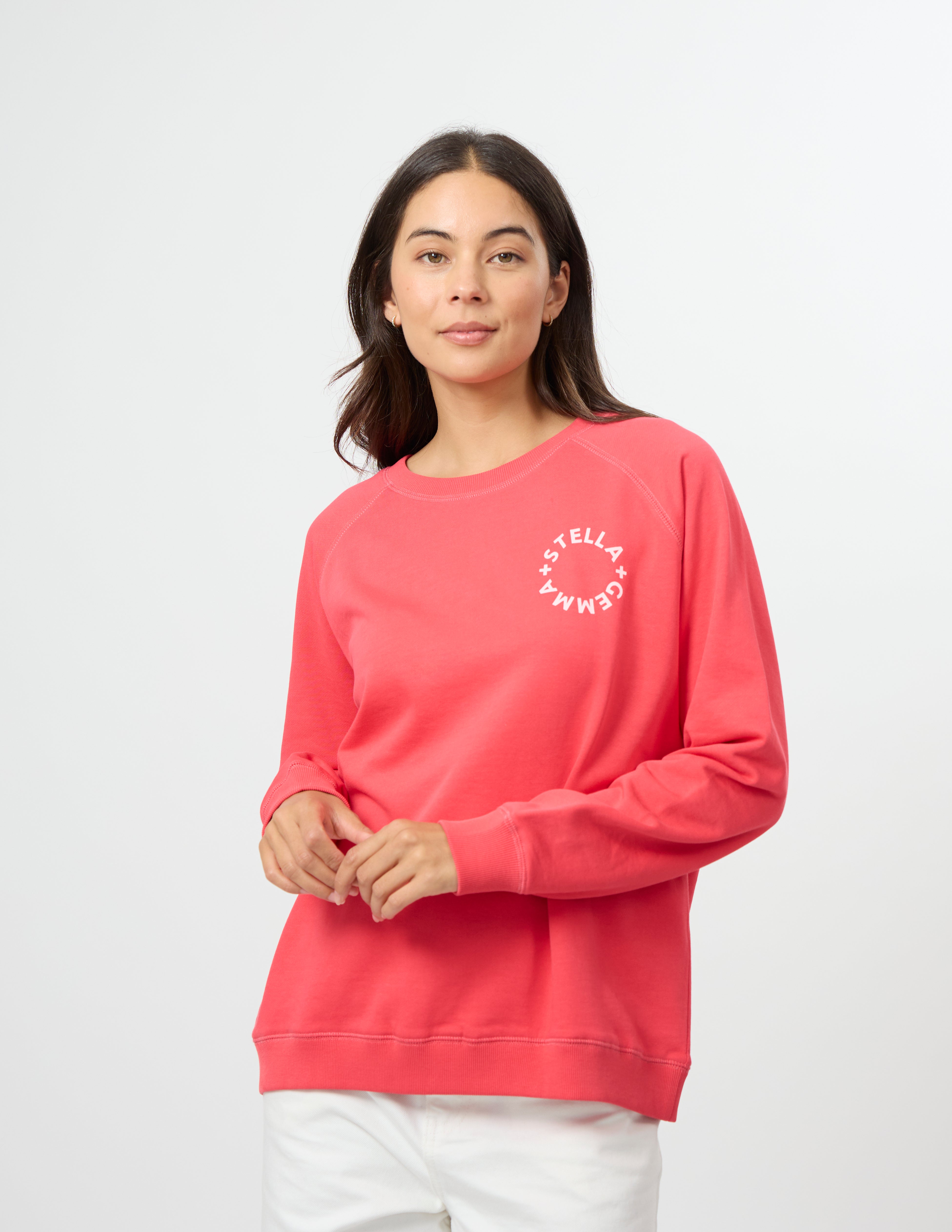 A woman wearing a coral pink crewneck sweatshirt with a white circular 'BELLA CARMEN' logo on the left chest, paired with white pants, against a light gray studio background.