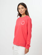 A woman modeling a coral pink crewneck sweatshirt with a white circular 'GEMMELTELL' logo on the left chest, paired with white pants, against a plain white studio background.