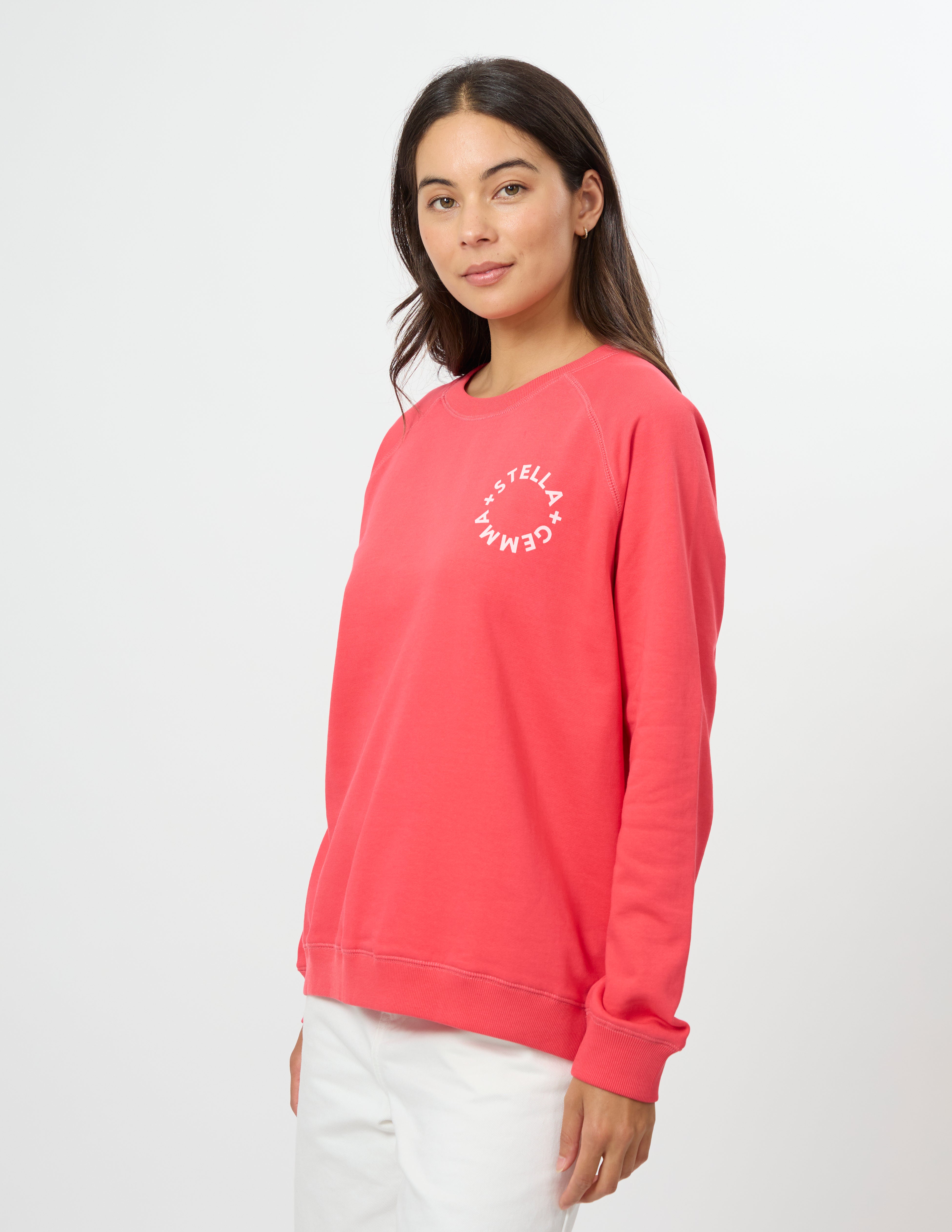 A woman modeling a coral pink crewneck sweatshirt with a white circular 'GEMMELTELL' logo on the left chest, paired with white pants, against a plain white studio background.