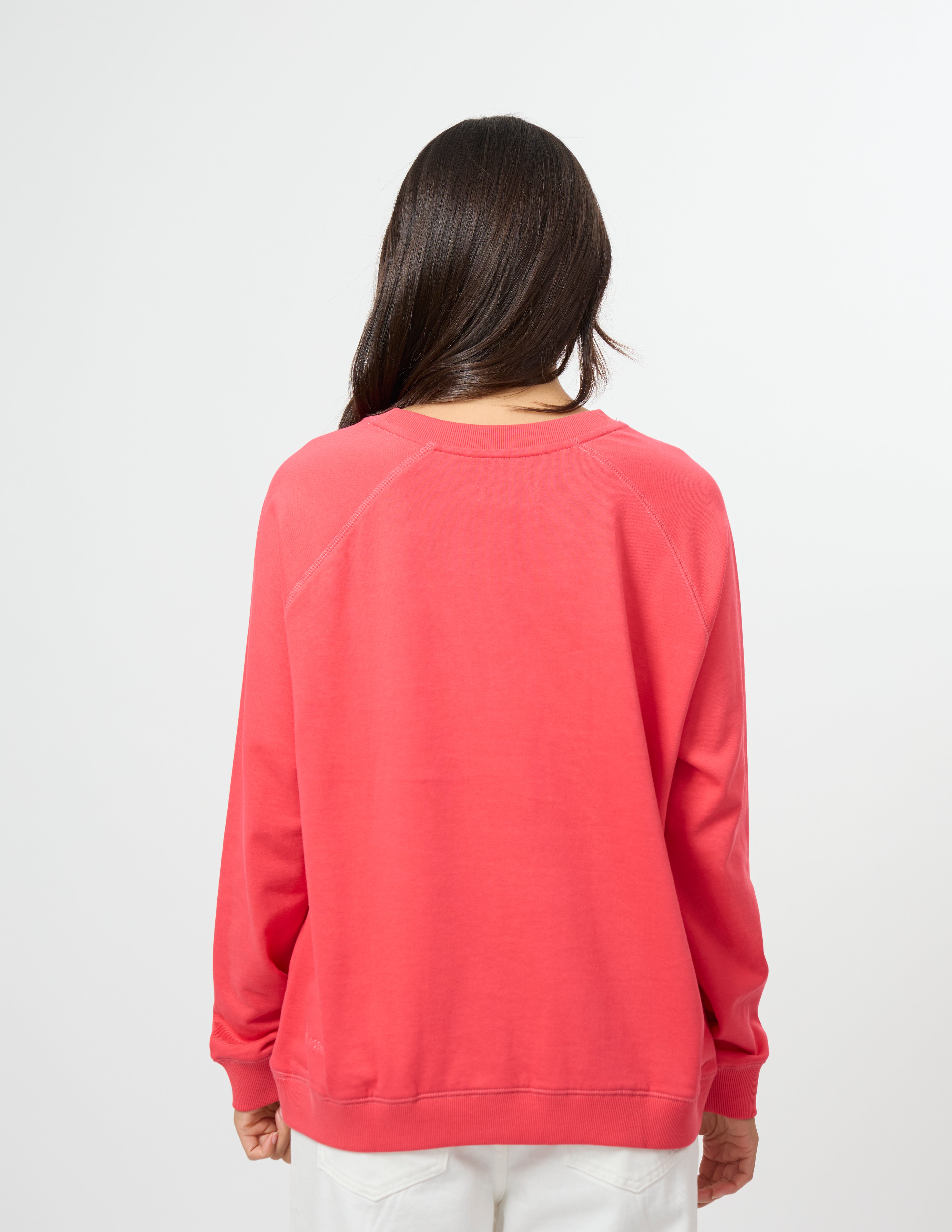 A woman viewed from behind wearing a coral pink crewneck sweatshirt and white pants, set against a plain light gray studio background.