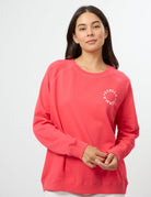 A woman wearing a coral pink crewneck sweatshirt with a white circular graphic on the left chest, photographed against a clean white studio background.