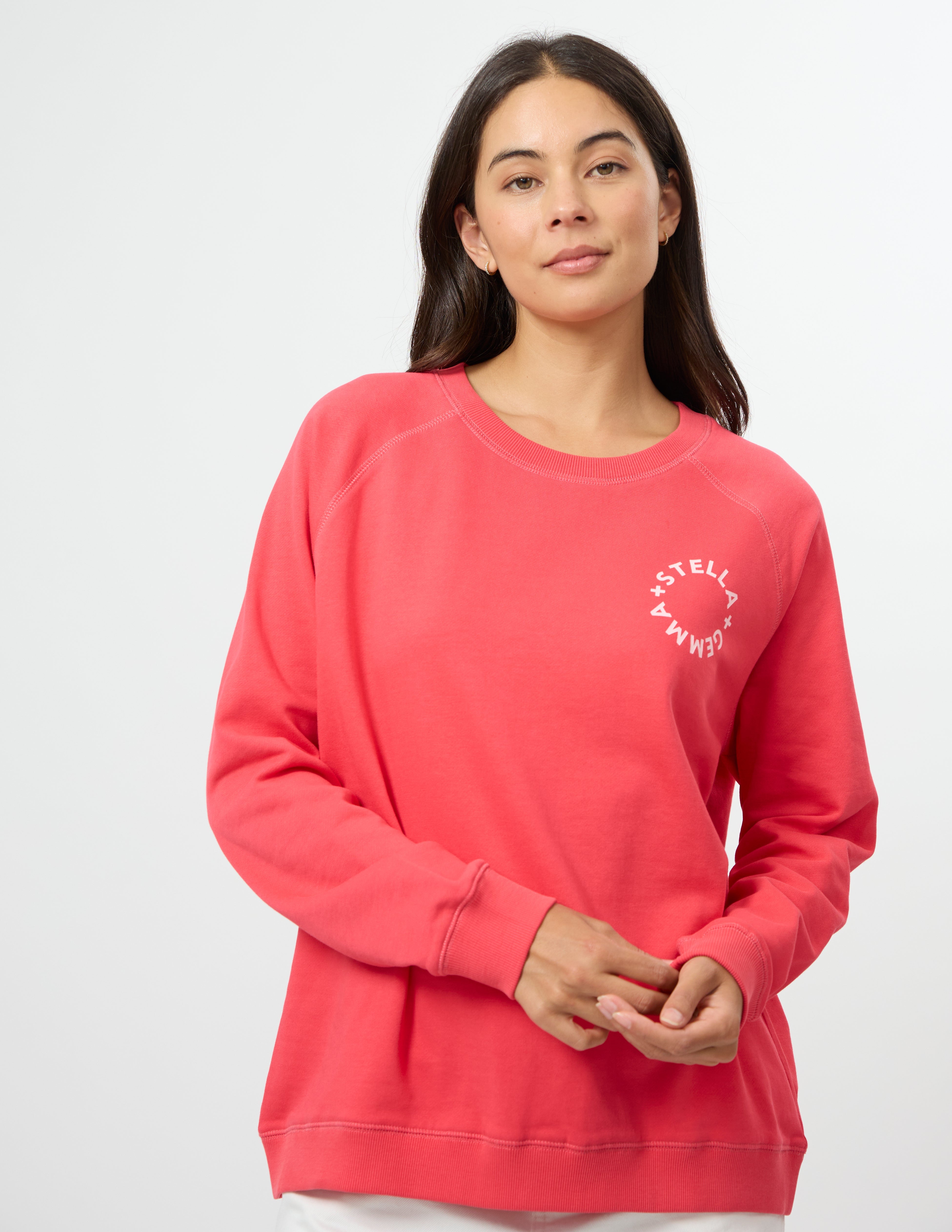 A woman wearing a coral pink crewneck sweatshirt with a white circular graphic on the left chest, photographed against a clean white studio background.