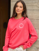 A woman wearing a coral pink crewneck sweatshirt with a white circular 'STELLA GEMMA' logo on the chest, standing against a rustic wooden wall.