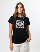 A woman modeling a black short-sleeve t-shirt with a white square mandala graphic on the chest, paired with off-white shorts, against a plain white studio background.