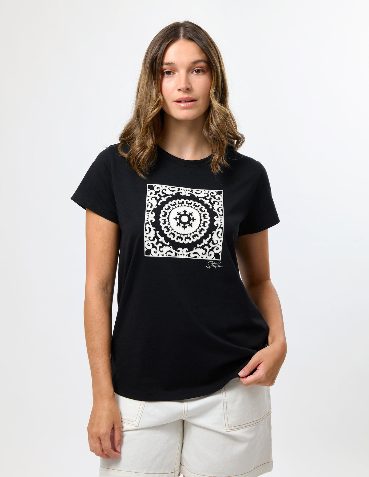A woman modeling a black short-sleeve t-shirt with a white square mandala graphic on the chest, paired with off-white shorts, against a plain white studio background.