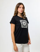 A woman modeling a black short-sleeve t-shirt with a white square mandala graphic on the chest, paired with white shorts, against a plain light gray background.