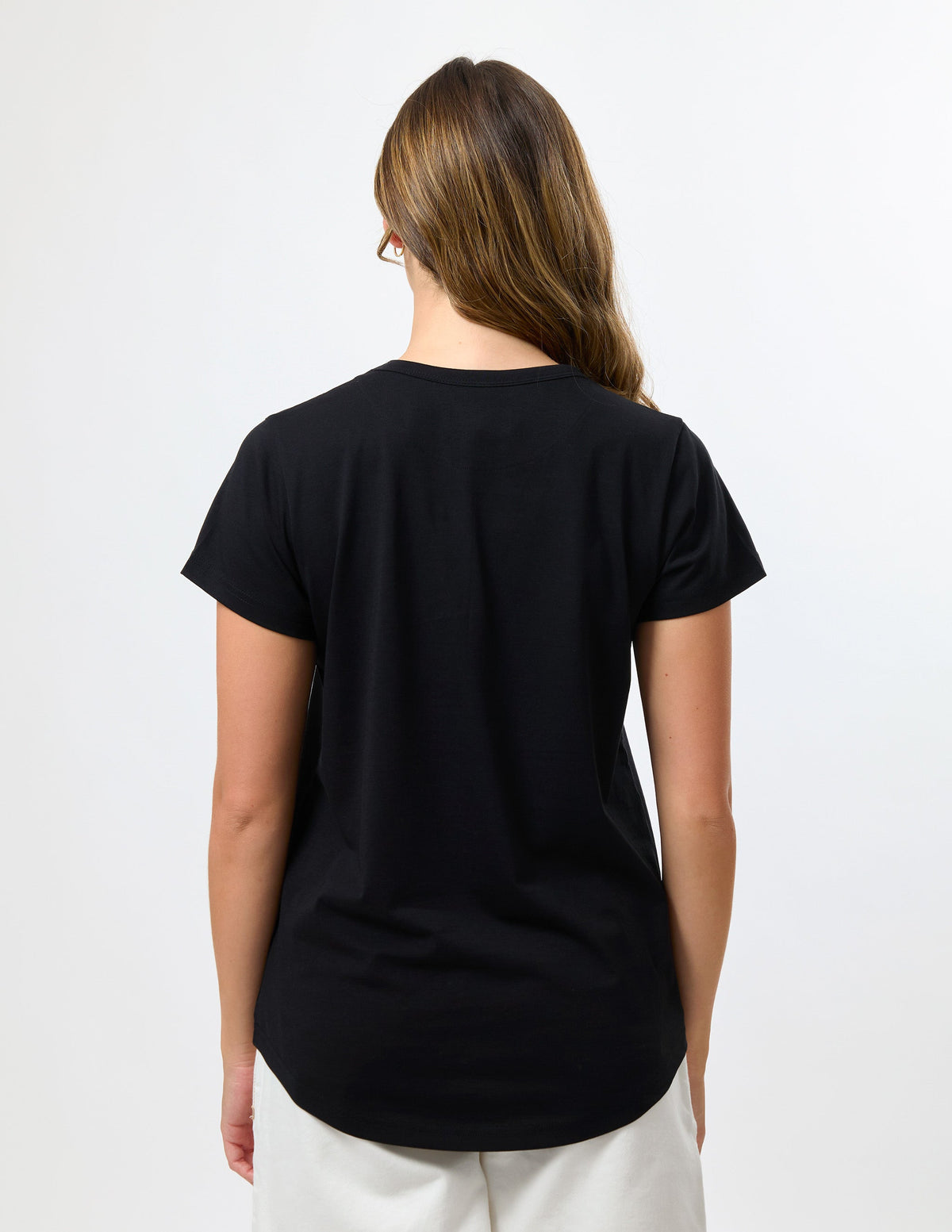 A woman viewed from the back wearing a plain black short-sleeve t-shirt and off-white pants against a clean white studio background.