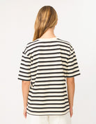 A woman viewed from the back wearing a short-sleeve striped t-shirt with horizontal black and cream stripes, paired with cream pants, against a plain white background.
