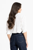 A woman shown from the back wearing a white textured blouse with gathered ruffles and dark blue jeans, photographed against a clean white studio background.