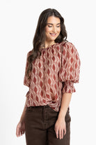 A woman wearing a rust-brown and cream wavy patterned blouse with puff sleeves, paired with dark brown pants, against a plain white background.