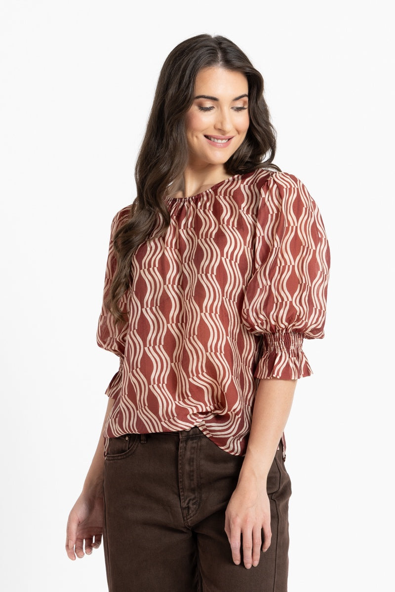 A woman wearing a rust-brown and cream wavy patterned blouse with puff sleeves, paired with dark brown pants, against a plain white background.