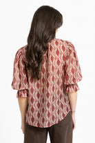 A woman viewed from the back wearing a rust-red and cream wavy patterned blouse with puff sleeves, paired with brown pants, against a plain white background.