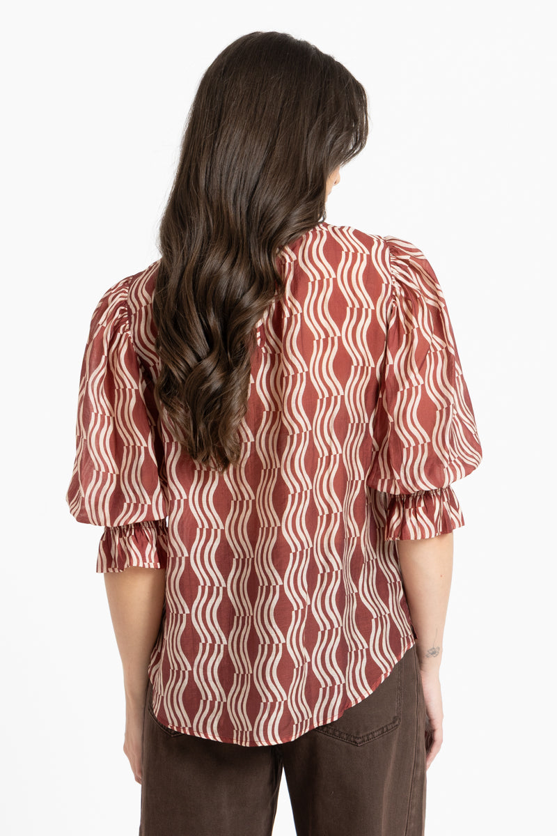 A woman viewed from the back wearing a rust-red and cream wavy patterned blouse with puff sleeves, paired with brown pants, against a plain white background.