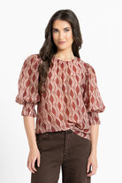 A woman modeling a rust-red and cream wavy patterned blouse with puff sleeves and a gathered waist, paired with dark brown pants, against a plain white background.