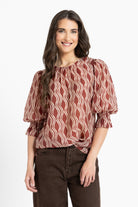 A woman modeling a rust-brown and cream wavy patterned blouse with puff sleeves, paired with dark brown pants, against a plain white background.