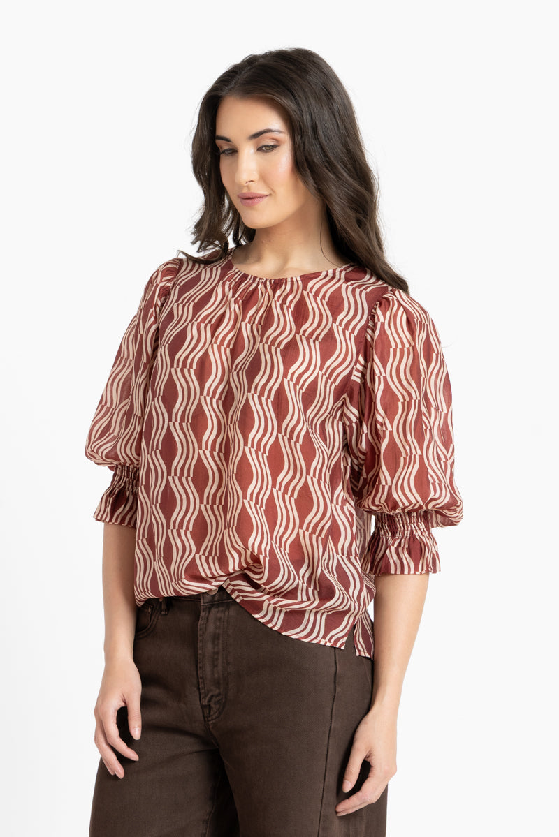 A woman wearing a rust-brown and white wavy patterned blouse with puff sleeves and dark brown pants, standing against a plain white studio background.