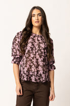 A woman modeling a dark brown blouse with a pink floral pattern and puff sleeves, paired with brown pants, against a plain off-white background.