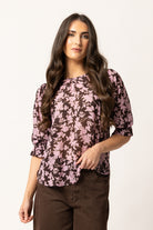 A woman modeling a three-quarter sleeve blouse with a pink and dark brown floral pattern, paired with brown pants, against a plain off-white background.