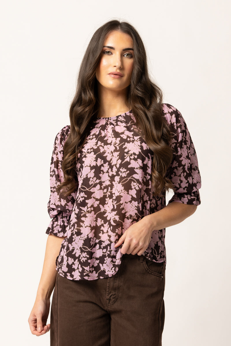 A woman modeling a three-quarter sleeve blouse with a pink and dark brown floral pattern, paired with brown pants, against a plain off-white background.
