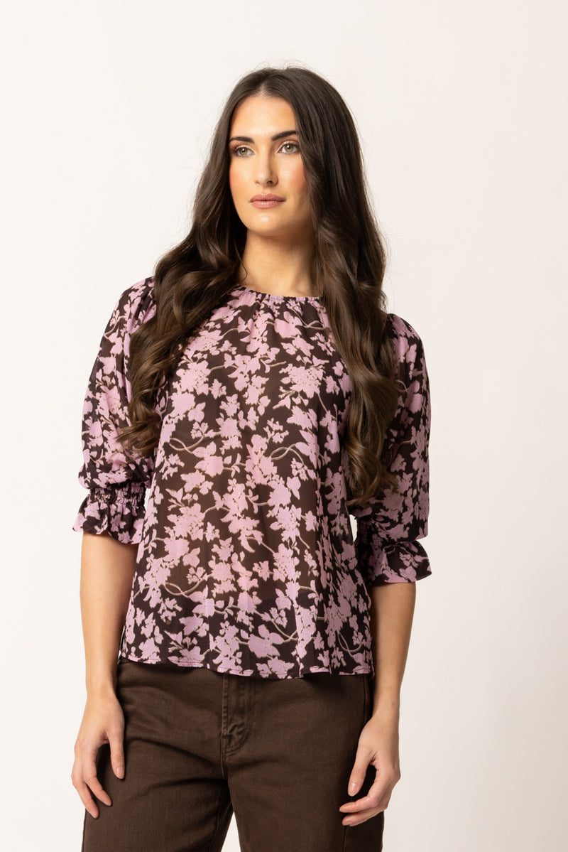 A woman modeling a dark brown long-sleeve blouse with a pink floral pattern, paired with dark brown pants, against a plain off-white background.