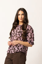 A woman modeling a short-sleeve blouse with a pink and dark brown floral pattern, paired with brown pants, against a plain off-white background.