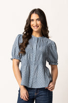 A woman modeling a light blue-gray smocked blouse with short puff sleeves and a subtle geometric pattern, paired with dark blue jeans against a plain white studio background.