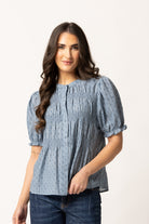 A woman modeling a light blue smocked blouse with short puff sleeves and a subtle geometric pattern, paired with dark blue jeans against a plain white studio background.