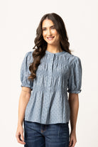 A woman modeling a light blue patterned blouse with puff sleeves and a smocked bodice, paired with dark blue jeans, against a plain white studio background.