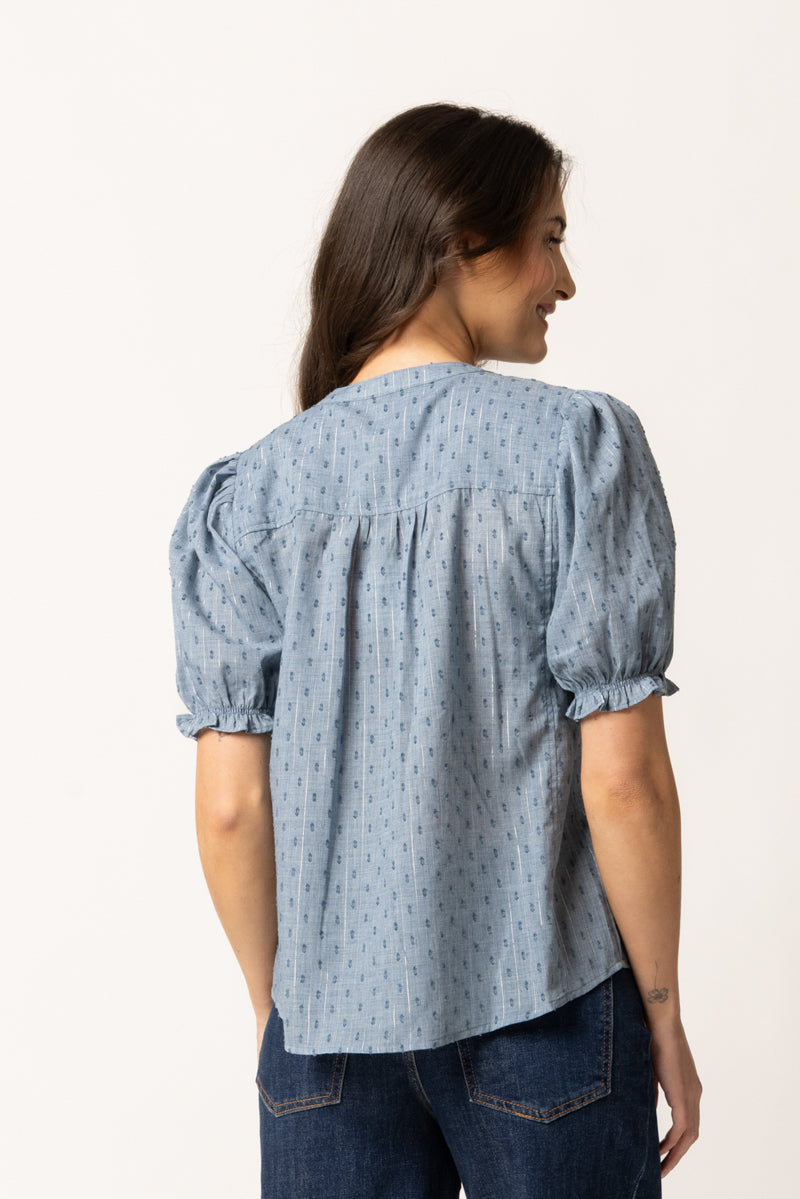 A woman shown from the back wearing a light blue patterned blouse with puff sleeves and dark blue jeans, photographed against a plain off-white studio background.
