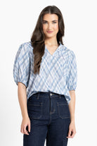 A woman modeling a light blue and white plaid short-sleeve blouse with puff sleeves and a V-neckline, paired with dark blue jeans, against a plain white studio background.